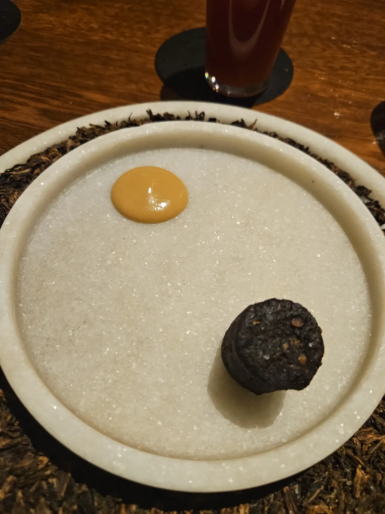 A small dark charred bite and a dollop of orange sauce on a plate over sugar-like sand