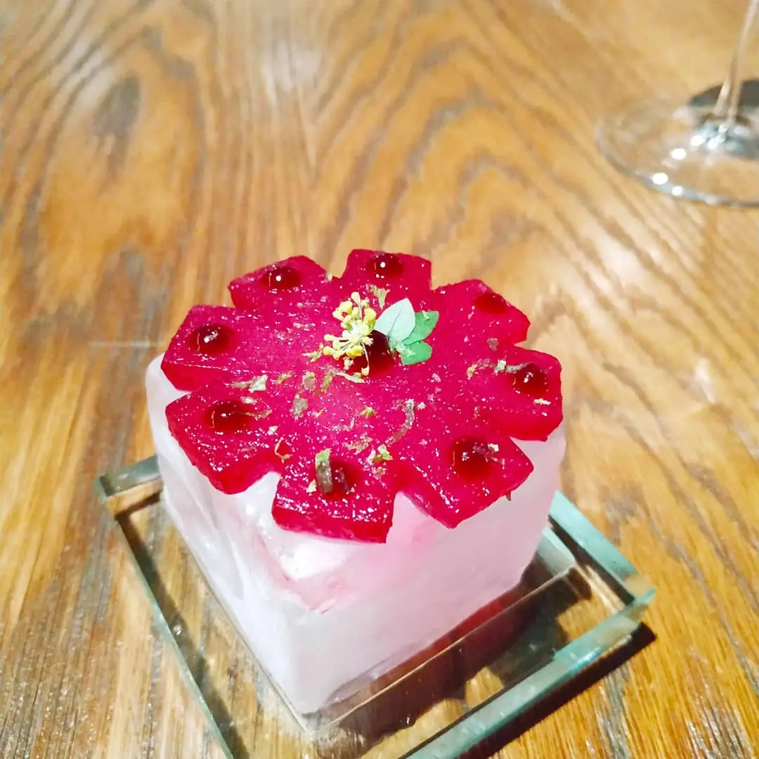 Square dessert with a flower-shaped red fruit topping and edible flower on a glass block