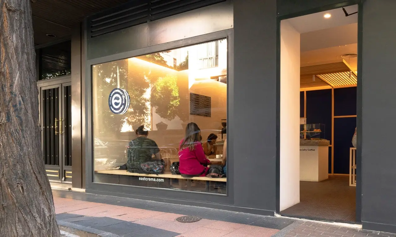 Street-level storefront of east crema coffee with a large window, logo decal and customers seated at a window bench