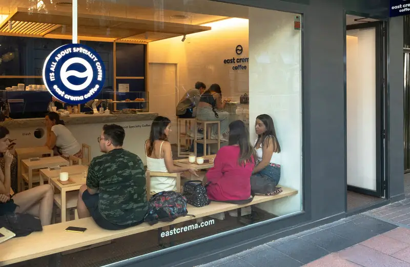 View through the window into east crema coffee showing the counter, blue logo sign and customers at wooden tables