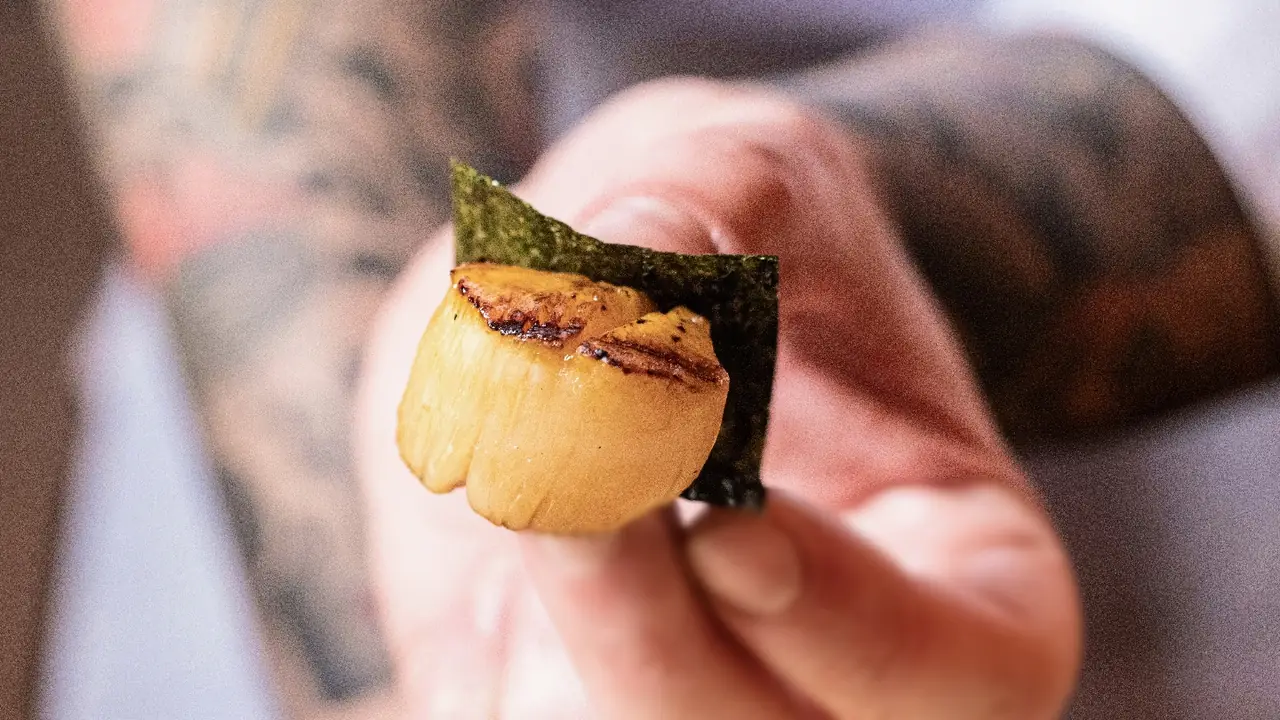 Hand holding a piece of seared scallop nigiri wrapped with nori