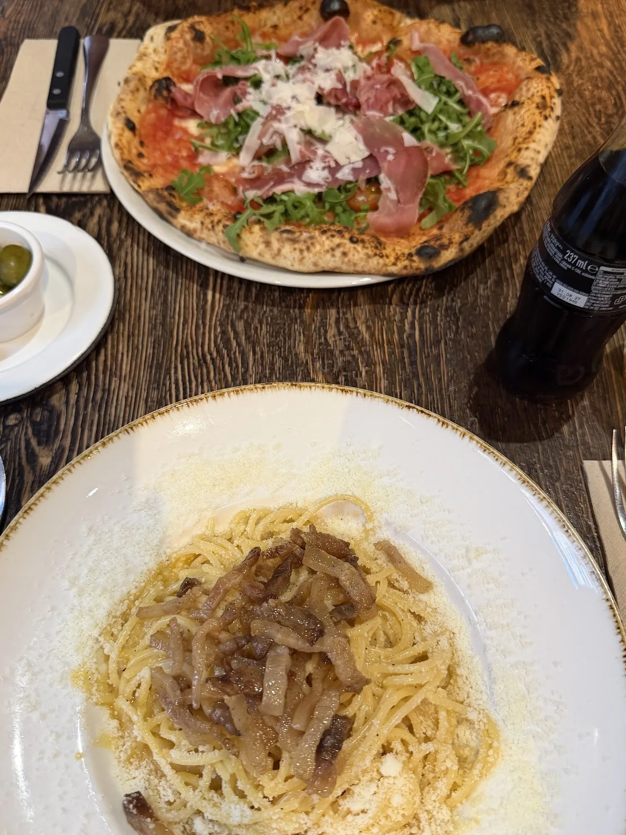 A plate of pasta with a pizza with cured ham on a wooden table