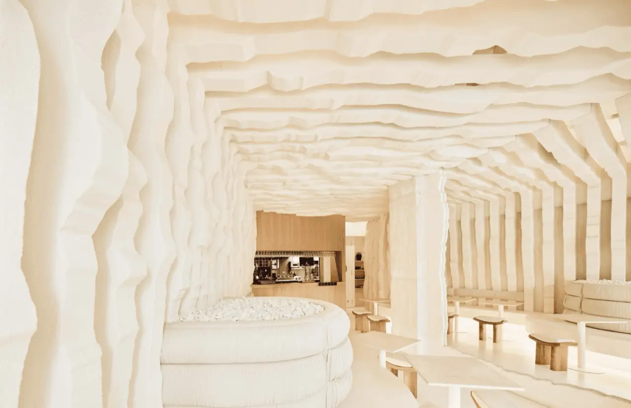 Dining room with sculpted cave-like cream-white ridged walls and ceiling, a curved white banquette holding white pebbles, timber tables and stools, and a bar in the background