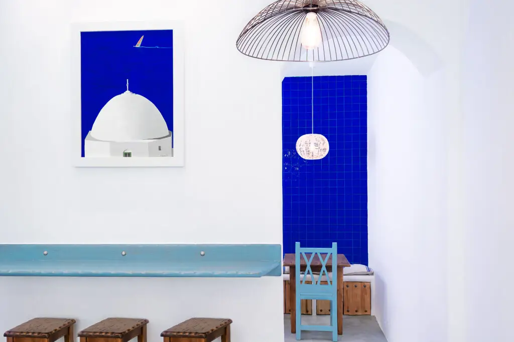 White-walled interior with a cobalt-blue tiled niche, framed Santorini-dome photograph, a blue-painted chair and wooden stools along a light-blue shelf