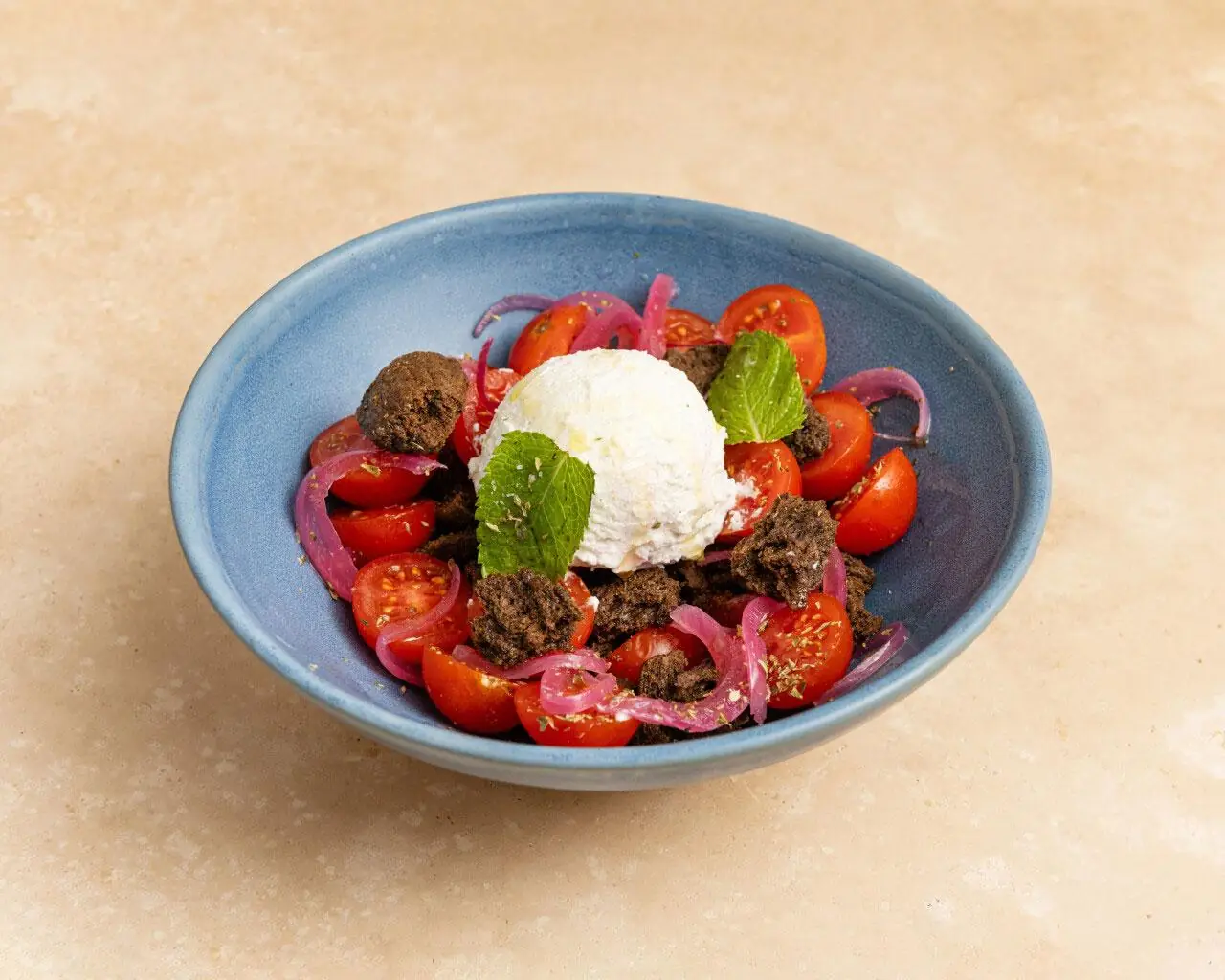 Halved cherry tomatoes with a scoop of soft cheese, red onion, olive crumble and mint in a blue ceramic bowl