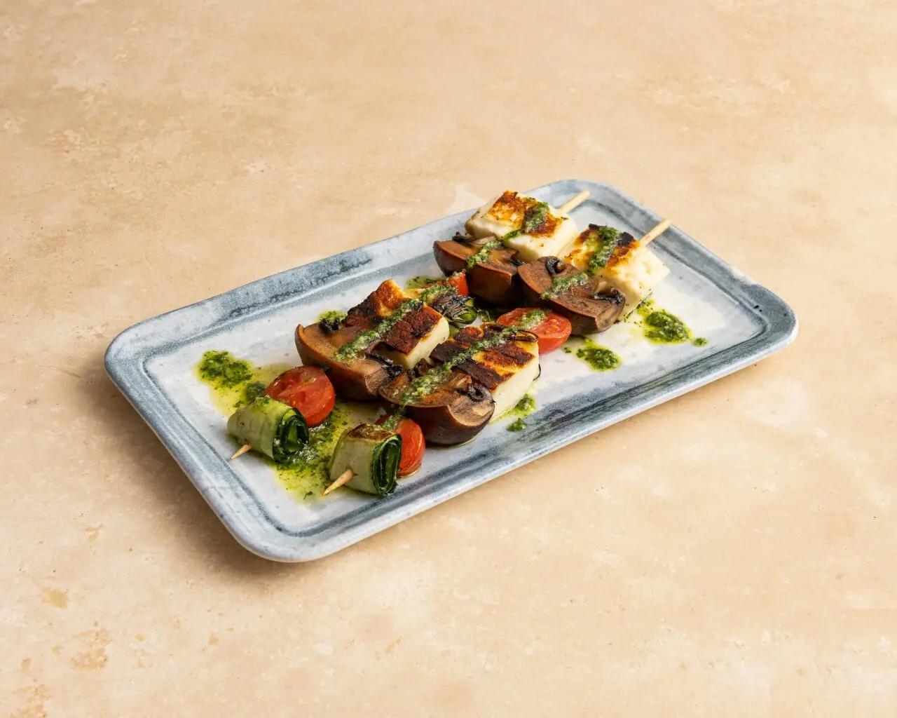 Grilled halloumi, mushroom, cherry tomato and courgette skewers drizzled with pesto on a rectangular ceramic plate