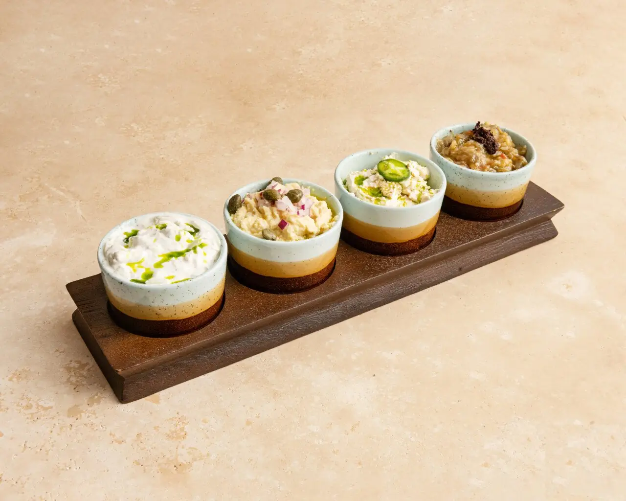 Wooden board holding four small ceramic bowls of Greek dips including tzatziki, hummus and eggplant