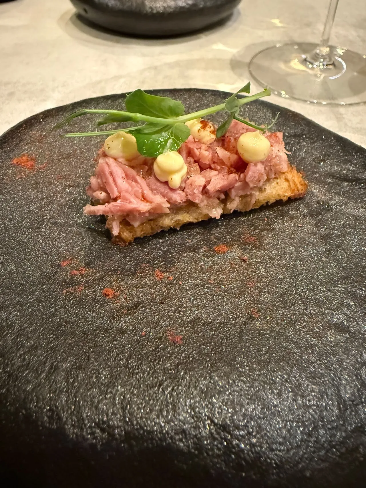 A meat or fish tartare on a dark plate with herb garnish and pickled onions