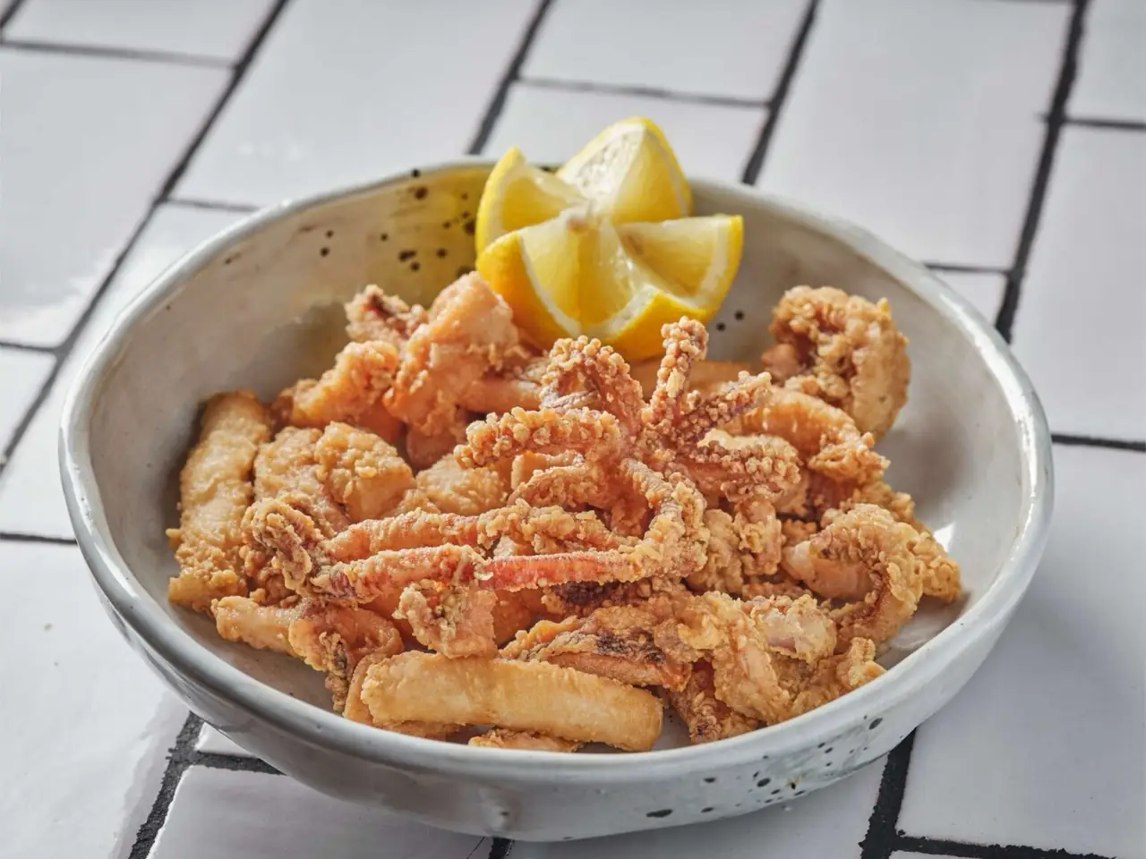 Fried calamari and battered seafood pieces in a stoneware bowl with lemon wedges, on white tile flooring