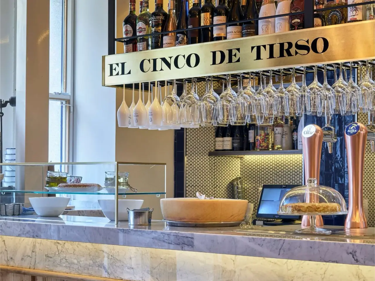 Bar interior with a brass 'El Cinco de Tirso' sign, hanging wine glasses, liquor bottles, beer taps, and a cheese wheel and cake under a glass dome on a marble counter