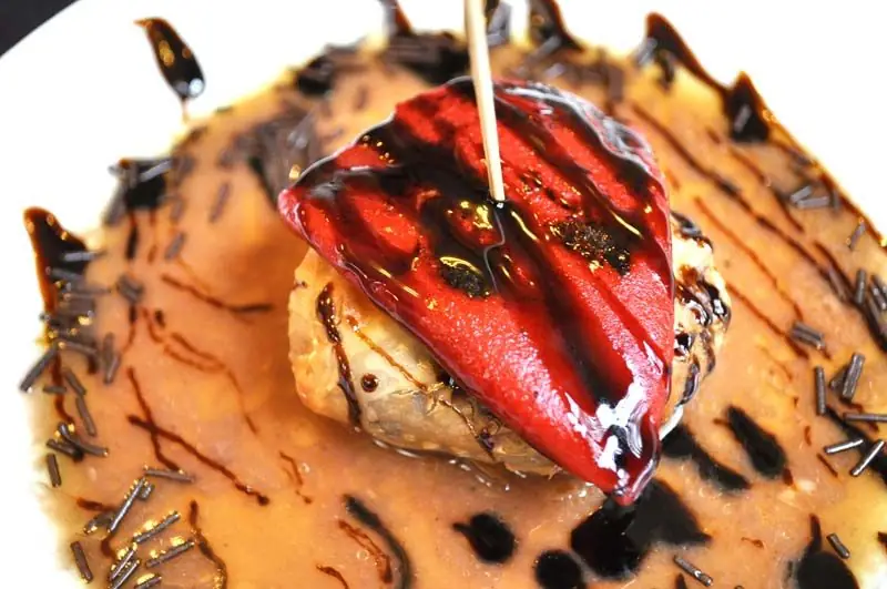 A roasted red piquillo pepper stuffed and glazed with dark balsamic, held by a wooden skewer over a pool of light sauce on a white plate