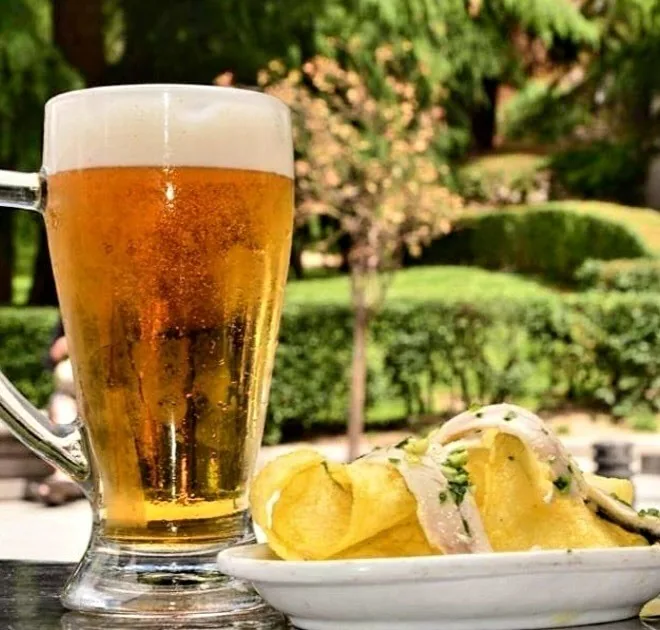 A tall glass of beer beside a plate of food with a garden behind