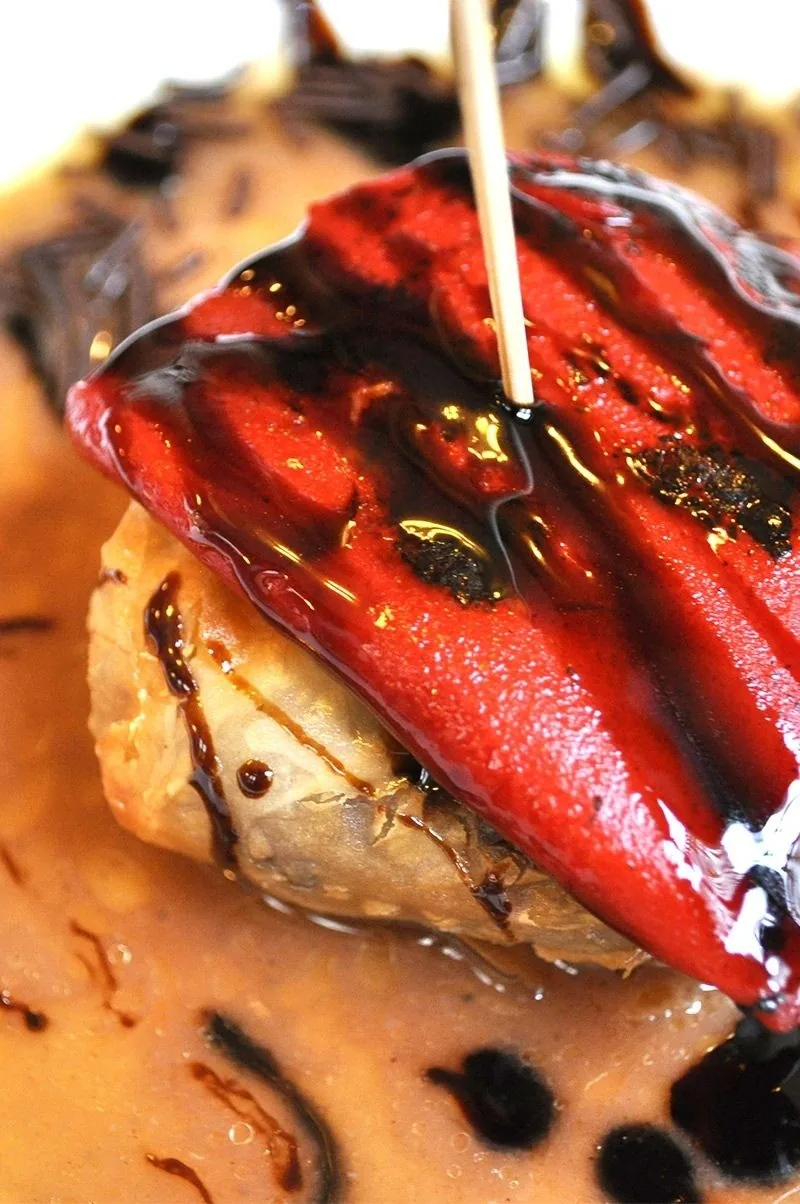 A skewered tapa topped with a glossy red pepper in a dark sauce