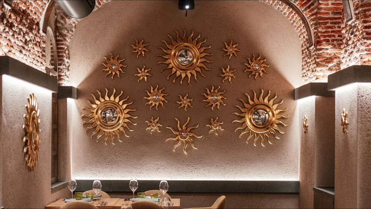 Dining room under a brick vaulted arch, its pink plaster back wall covered with a constellation of gilded sunburst mirrors, with laid tables and wine glasses in the foreground