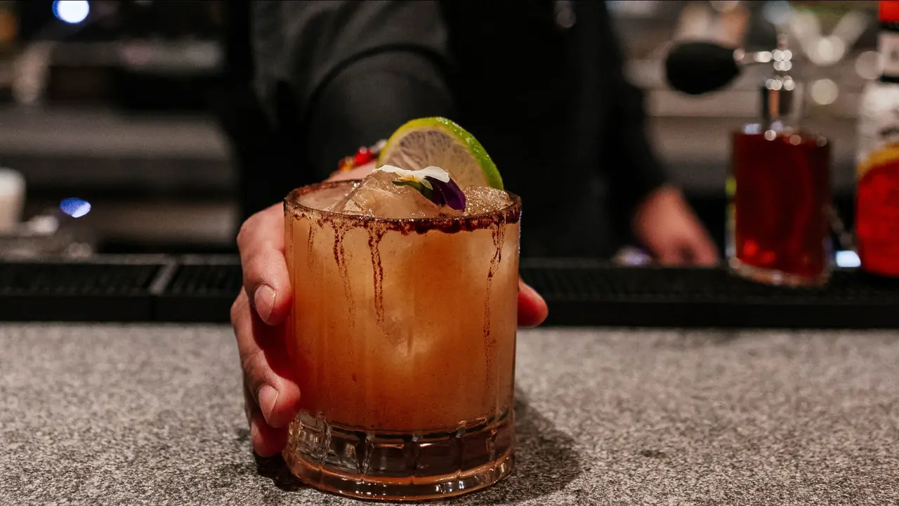 A hand sets down a rocks glass of pale orange cocktail over ice on a speckled stone bar, the rim crusted with dark red chilli salt that streaks down the glass, garnished with a lime wheel and a small white and purple flower