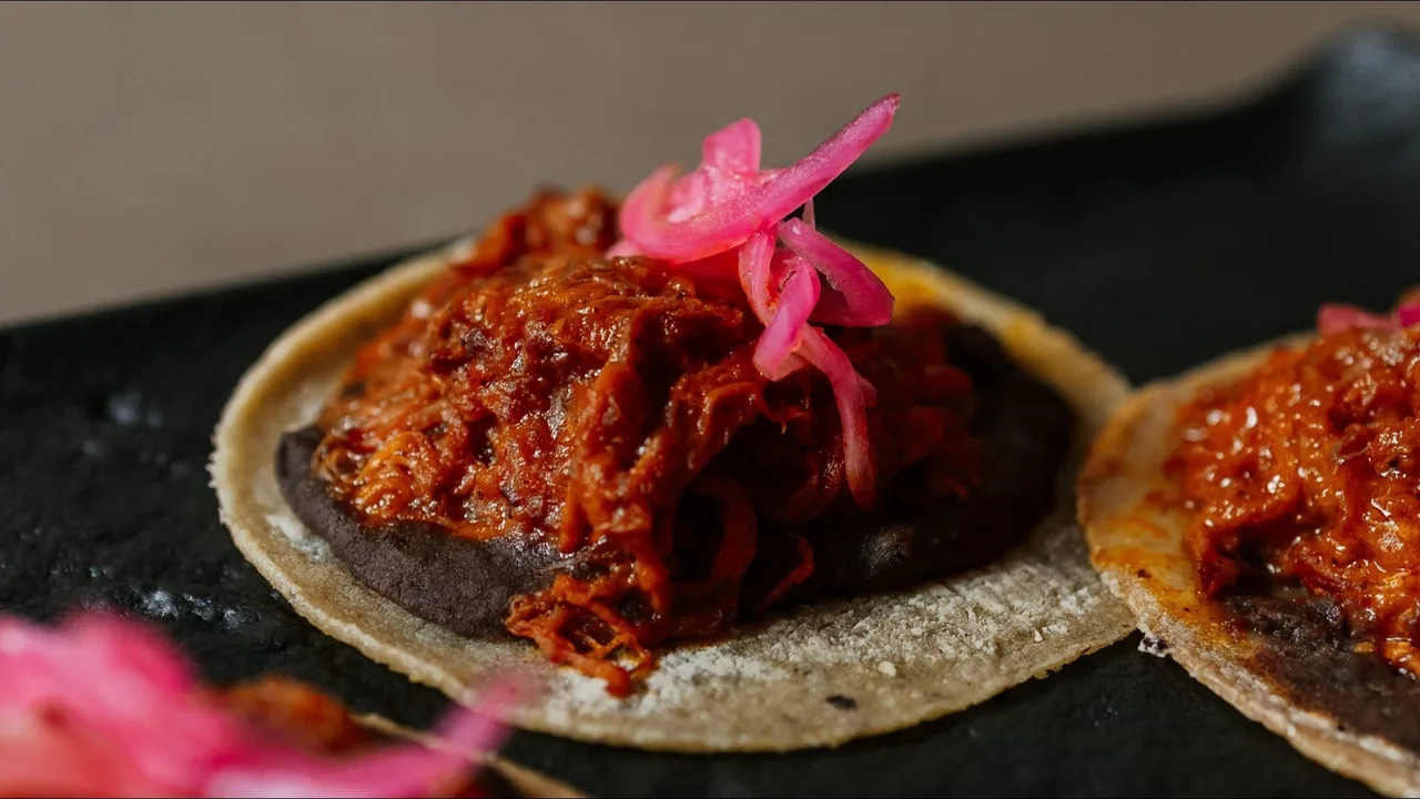 Two small tortillas topped with shredded red meat and pickled pink onion on a dark plate