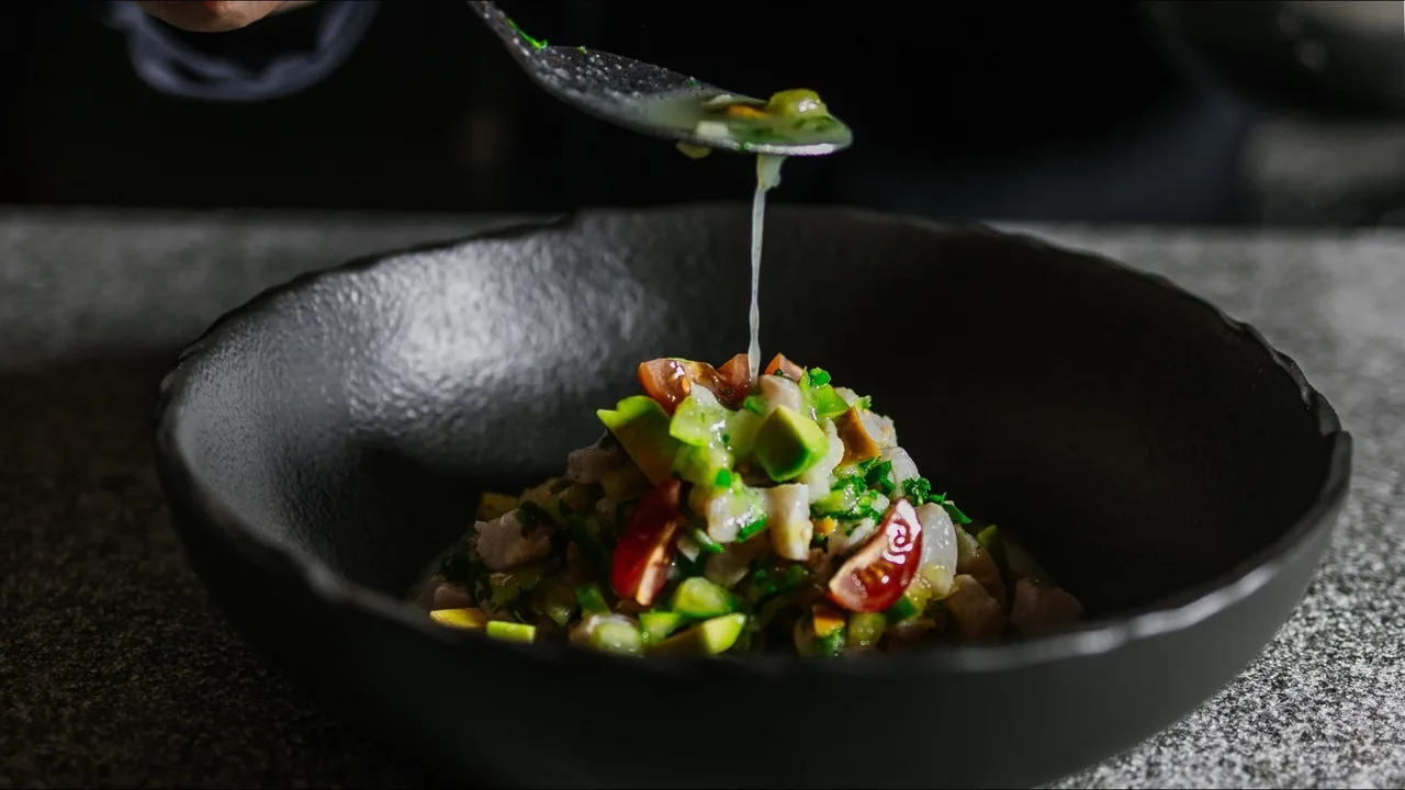 A black bowl of diced avocado, tomato and herbs with a spoon dripping green liquid over it
