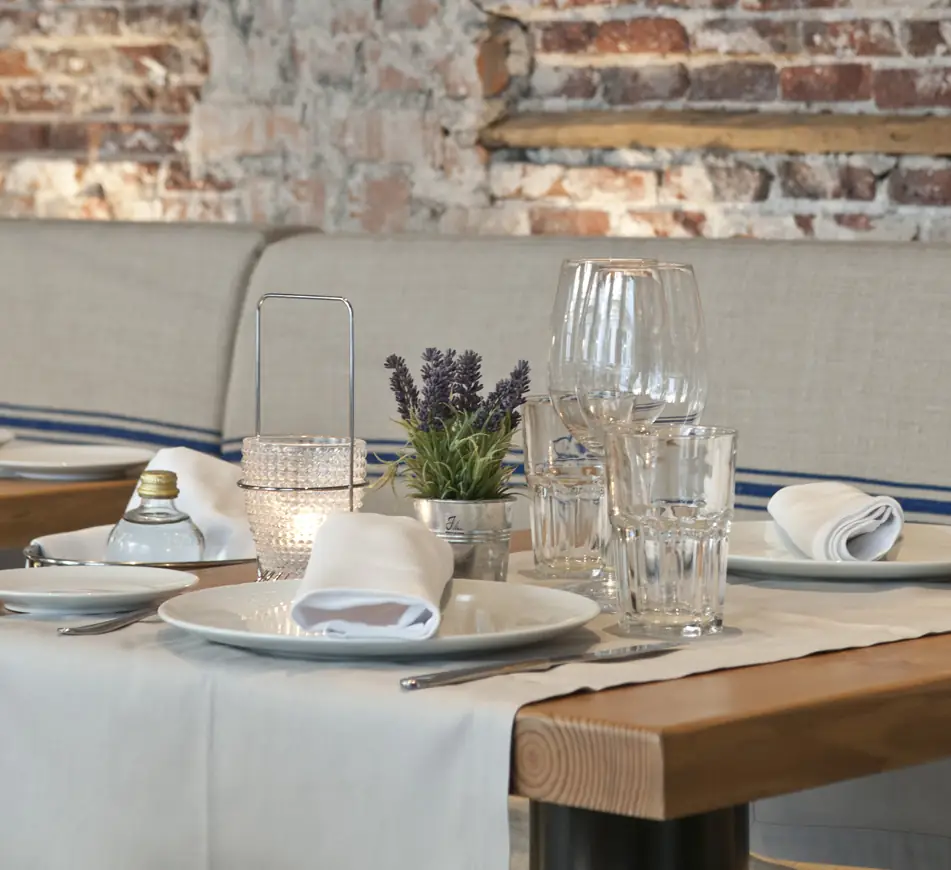 Set table with white plates, folded napkins, glassware and a small lavender pot against an exposed brick wall