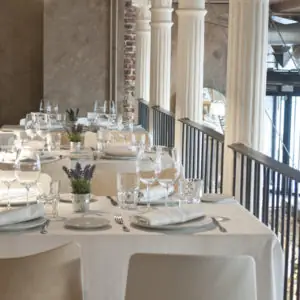 Dining room with a row of white-clothed set tables beside white columns and a railing
