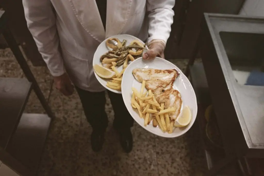 A waiter holds two white plates of food: grilled chicken breast with french fries and lemon, and fried squid with grilled green asparagus
