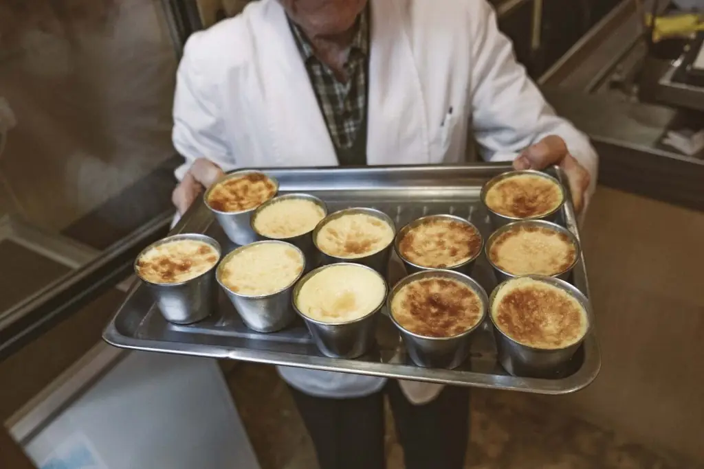 A metal tray holding ten individual metal cups of baked custard dessert with lightly browned tops, carried by a cook
