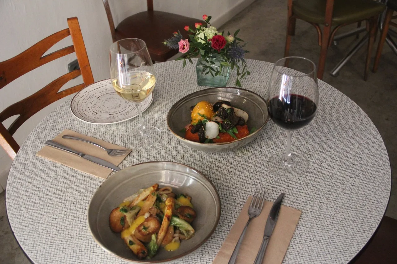 Round table with two mushroom dishes, one over a fried base with black trumpet mushrooms and egg yolk, plus glasses of white and red wine.
