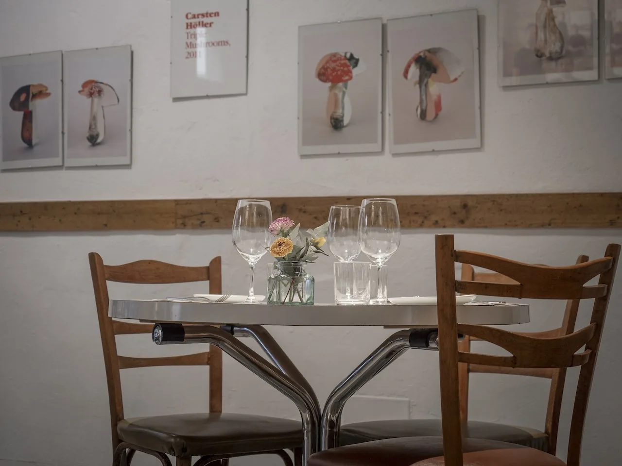 Small table set with glasses beneath a row of framed mushroom photographs on a whitewashed wall.
