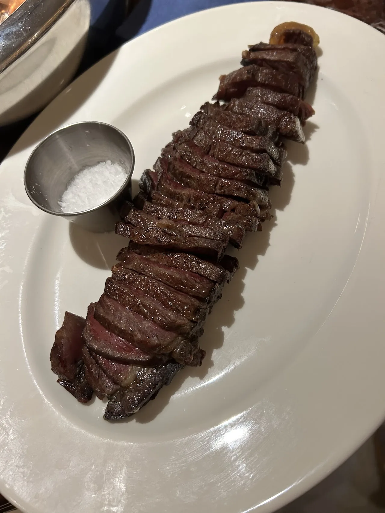 Sliced grilled steak arranged on a white plate with a small pot of salt
