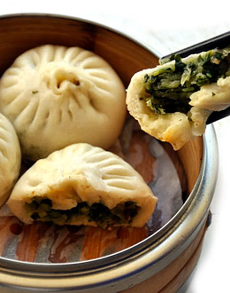 Steamed buns in a bamboo steamer, one torn open to show a bright green vegetable filling, with chopsticks lifting a piece clear