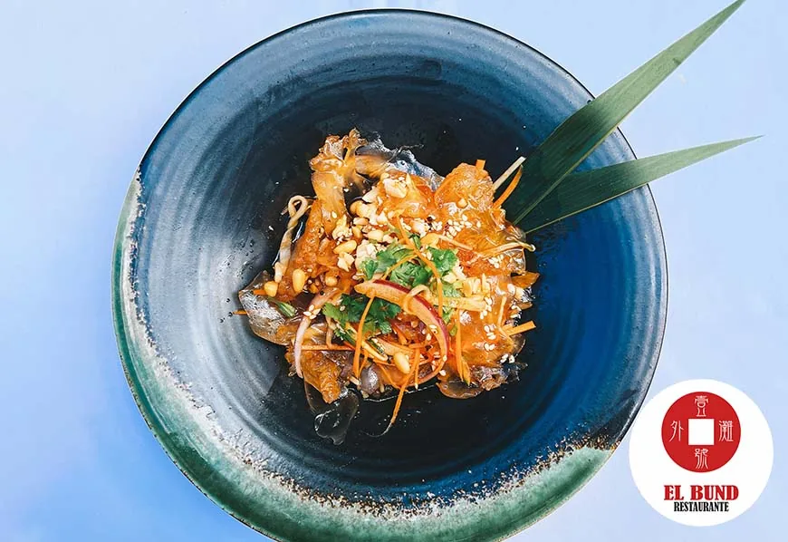 An overhead of a green-and-black glazed bowl of jellyfish salad with shredded carrot, red onion, coriander and crushed peanuts, a pandan leaf standing behind