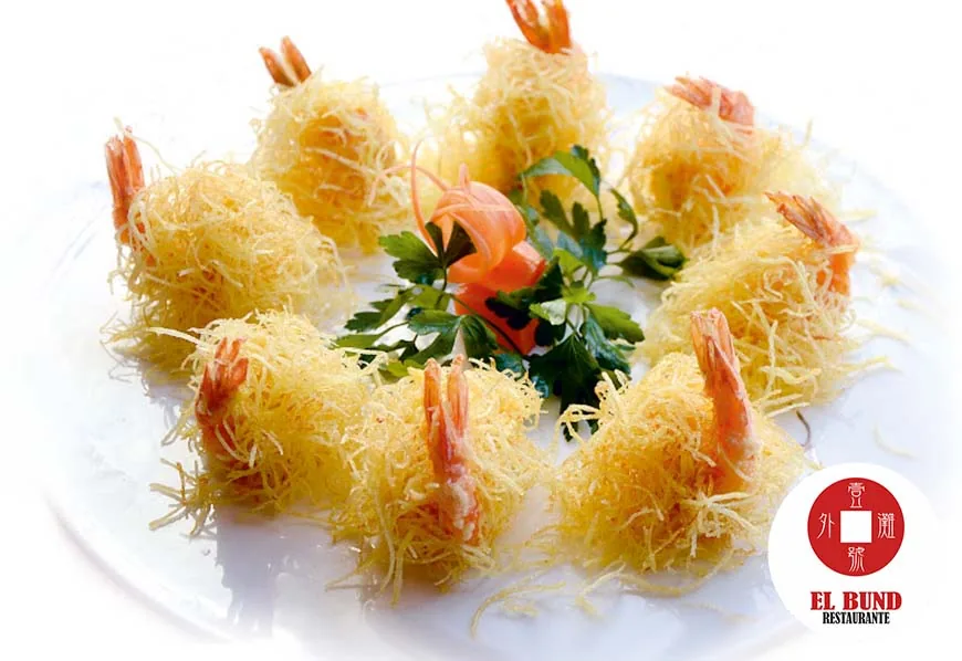Eight prawns wrapped in shredded crisp pastry arranged in a ring around a carved carrot flower and parsley on a white plate