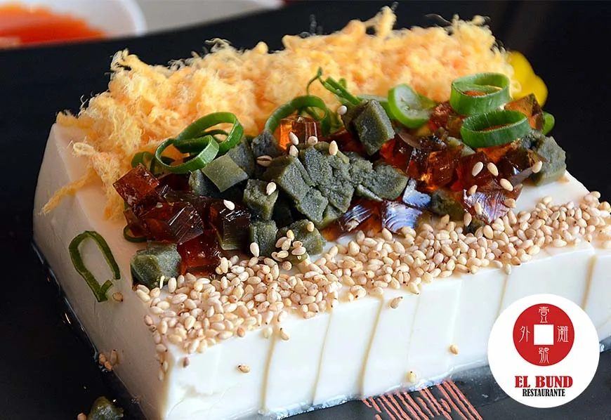 A block of silken tofu topped with century egg, pork floss, spring onion and sesame seeds on a dark plate