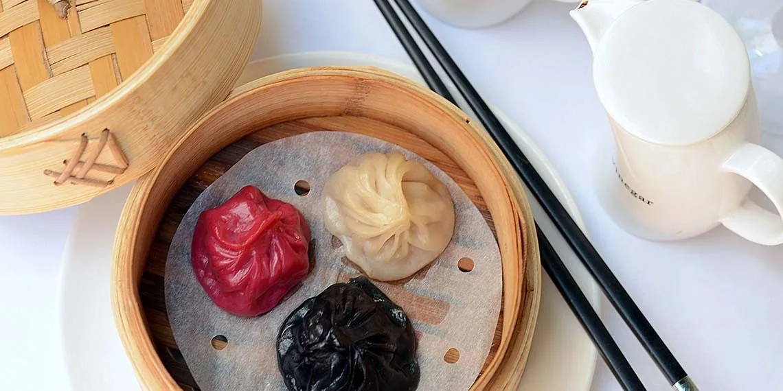 A bamboo steamer holding red, black and cream dumplings beside chopsticks