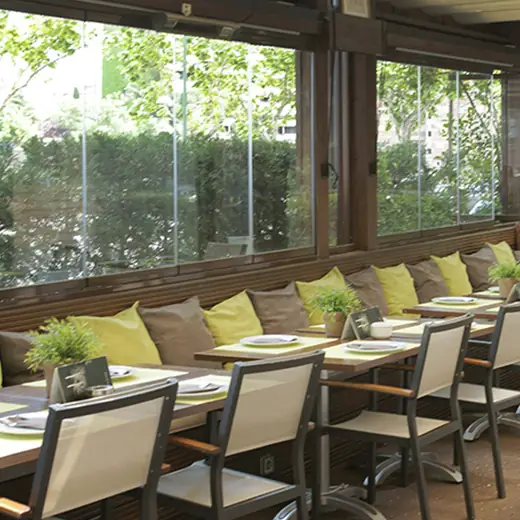 Glassed-in terrace dining area with a long banquette of green and mustard cushions, wooden tables set with green placemats, facing hedges outside.