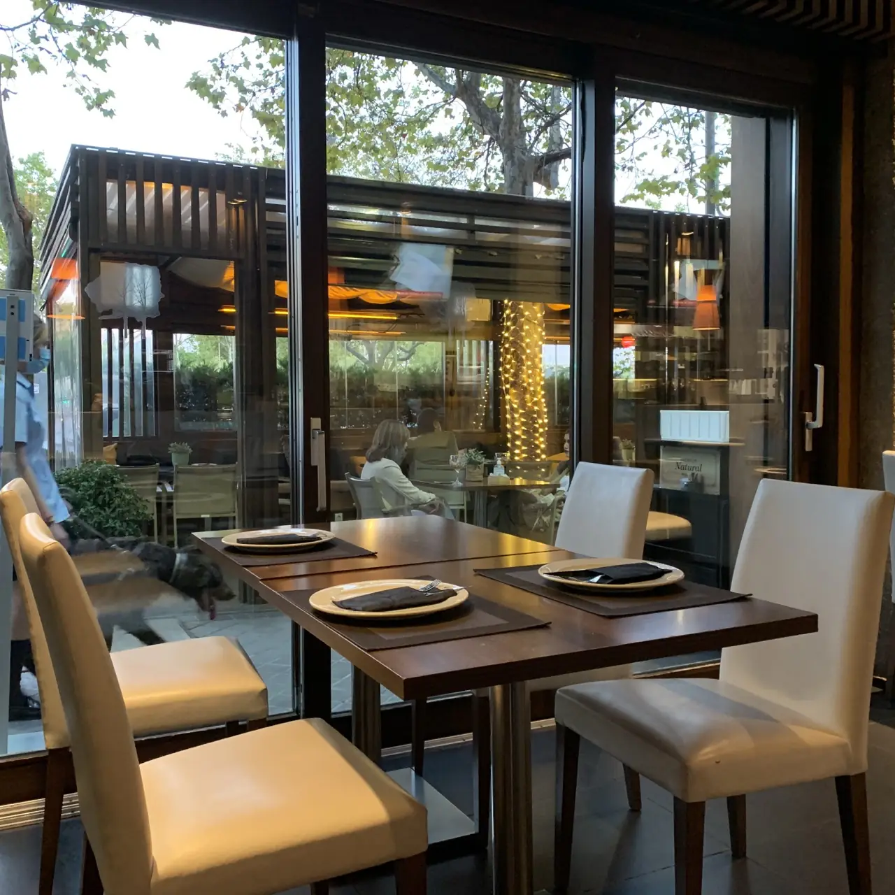 Table set for four with plates and dark napkins, seen through glass doors reflecting a wood-slatted covered terrace with string lights and outdoor seating.