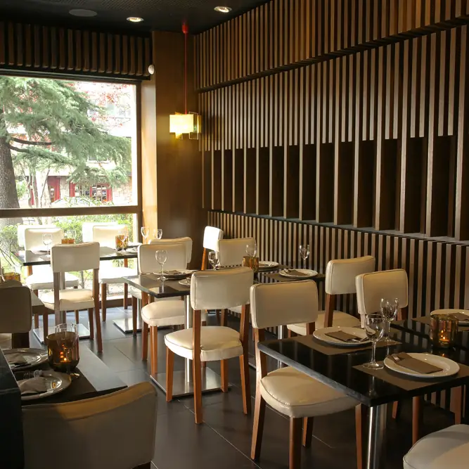 Dining room with dark wood slat wall paneling, cream chairs at black tables set with glassware, and a tree visible through the window.
