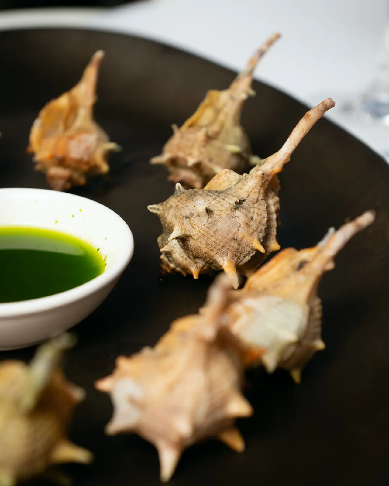 Cooked sea snails (cargols punxencs) served on a black plate with a small bowl of green herb oil for dipping, at El Camarote de Tomás