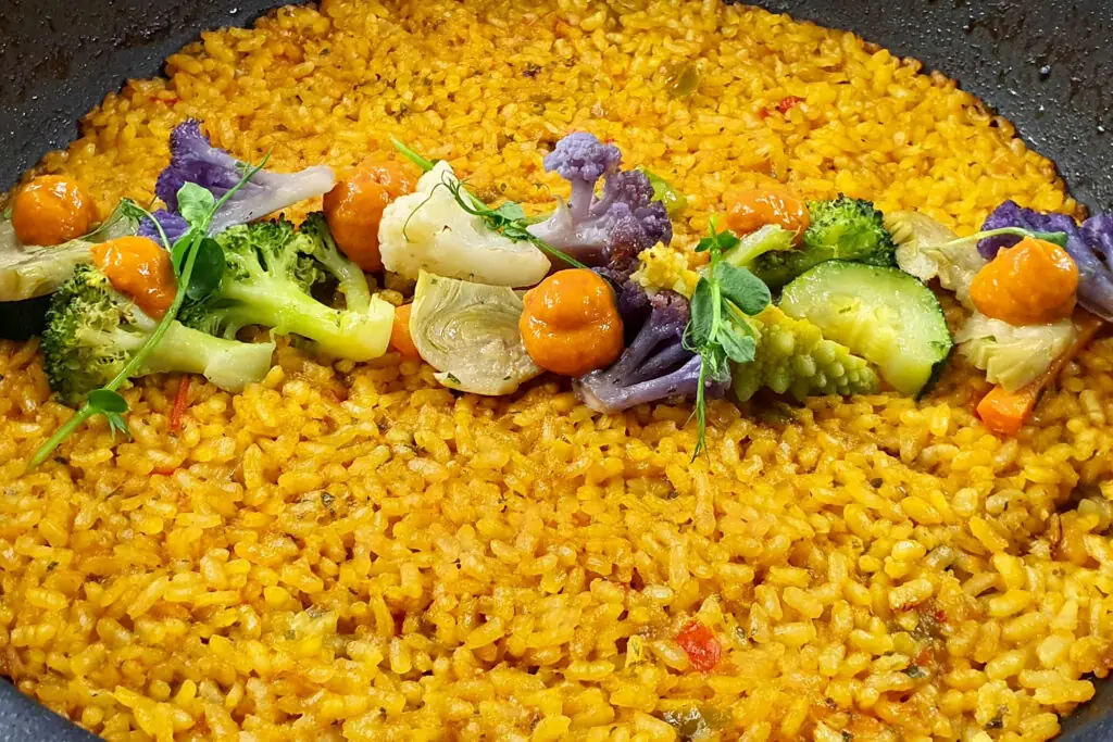 Saffron rice topped with purple cauliflower, broccoli, artichoke hearts, cucumber, and orange romesco-style dots, in a black paella pan, at El Cangrejo Loco