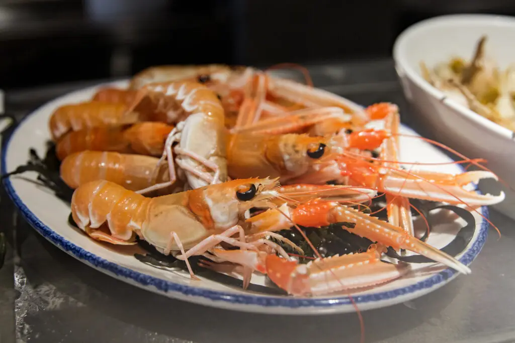 Whole cooked langoustines piled on a blue-rimmed plate over dark seaweed, with a bowl of food in the background, at El Cangrejo Loco