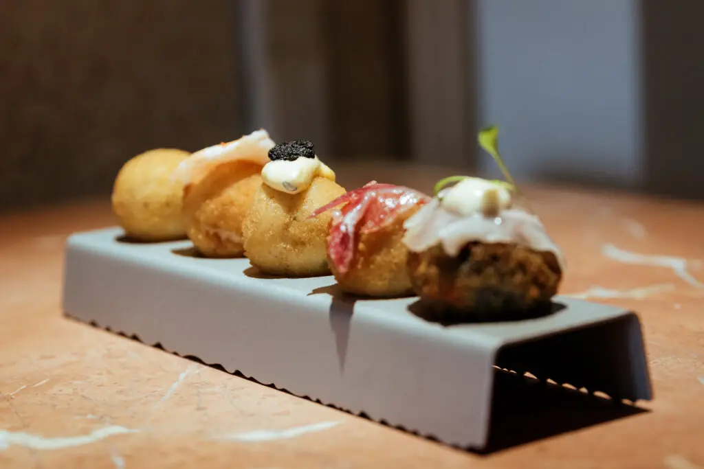 Five breaded croquettes and fritters lined up in a metal serving rack, topped with sauces, sliced meat, and a small herb sprig, at El Cangrejo Loco