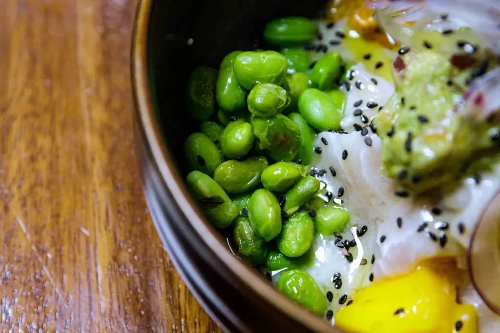 Close-up of a bowl with edamame beans, black sesame seeds, mango, and creamy sauce, on a wooden table, at El Cangrejo Loco
