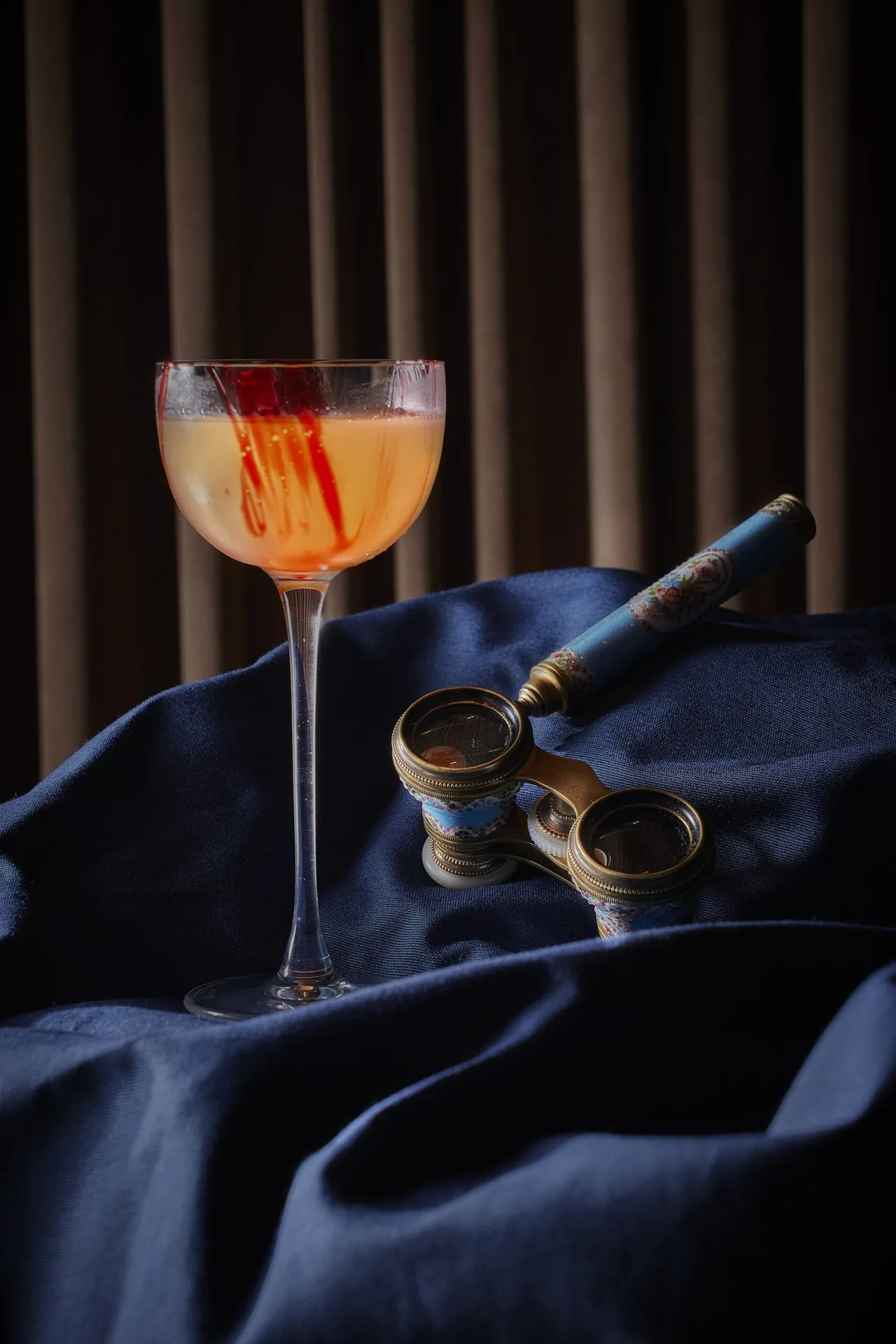 A coupe cocktail with a red garnish next to a small brass telescope on blue fabric