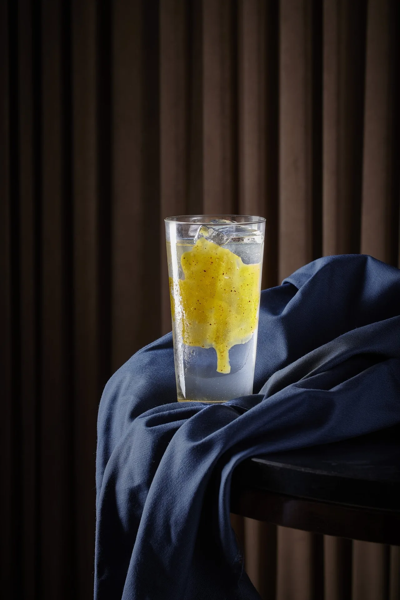 A tall glass with a yellow drink on blue fabric against brown curtains