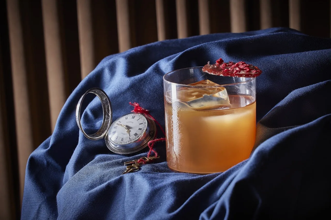 An amber cocktail garnished with berries on blue fabric