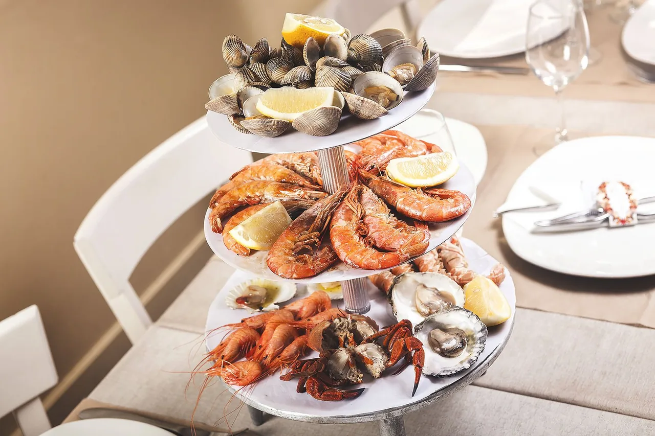 A three-tier seafood tower on a table: cockles and clams with lemon wedges on top, whole langostinos in the middle, and prawns, oysters, crab and langoustines on the base plate