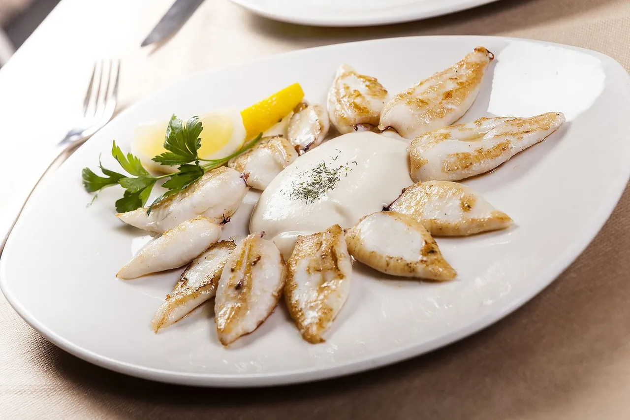 Grilled baby cuttlefish arranged in a ring around a pool of pale alioli dusted with dried herbs on a large white oval plate, with lemon wedges and a sprig of flat parsley to one side