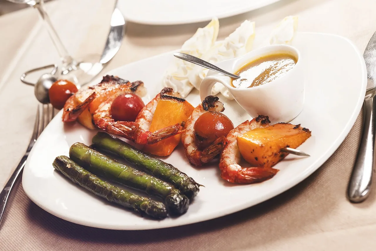 A skewer of grilled prawns with cherry tomatoes and squash on a white plate, with charred green asparagus, a fan of shaved white vegetable and a small jug of amber sauce