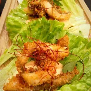 Crispy breaded chicken pieces in lettuce cups topped with creamy sauce and red chili threads on a wooden board