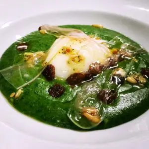 Poached egg over bright green vegetable puree with raisins, pine nuts and crisp tuiles on a white plate