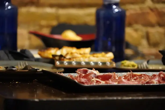 Sliced cured ham and salami on a black rectangular plate, with grilled flatbread and other tapas plates on the table behind, El Drapaire de la Cervesa Artesana.